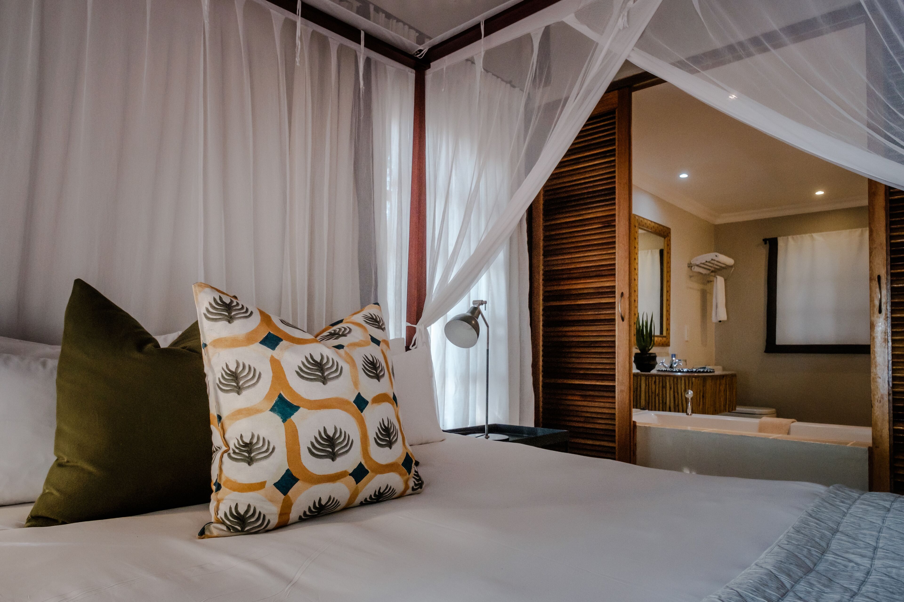 A bedroom with a bed, a nightstand, a lamp, and a bathroom visible through an open door.