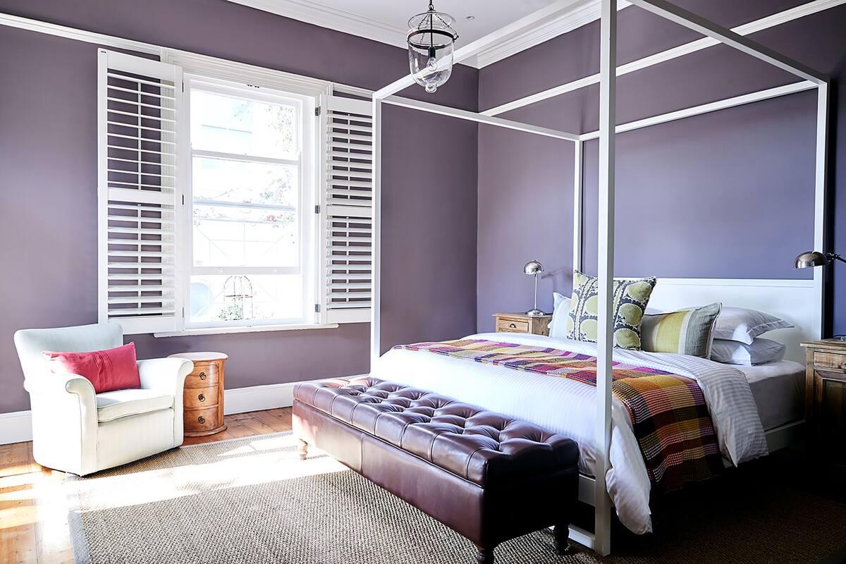 A bedroom with a four-poster bed, a chair, a bench, and a window with shutters.