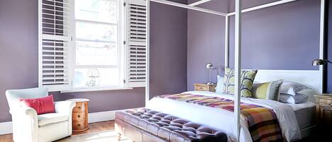 A bedroom with a four-poster bed, a chair, a bench, and a window with shutters.