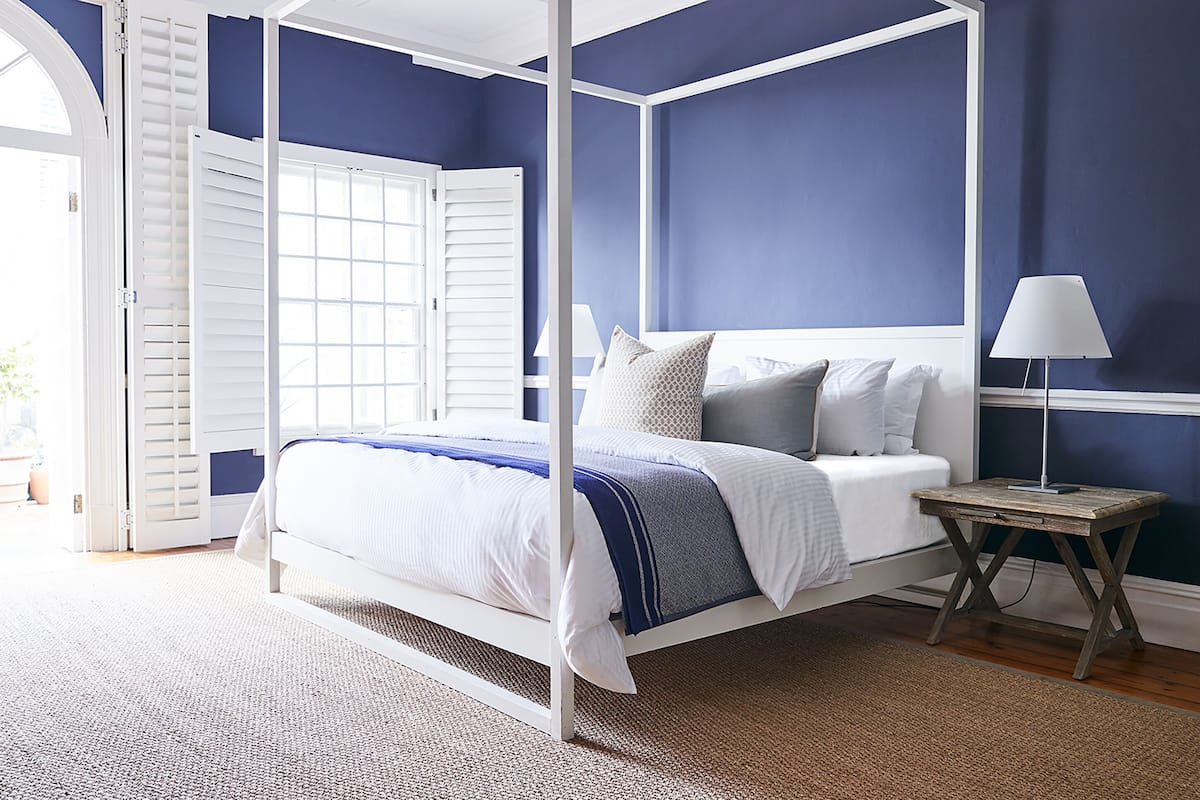 A four-poster bed with white bedding and a blue accent, a bedside table with a lamp, and a view of a door leading outside.