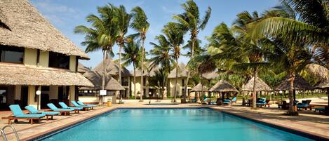 A swimming pool with lounge chairs, palm trees, and thatched-roof structures.