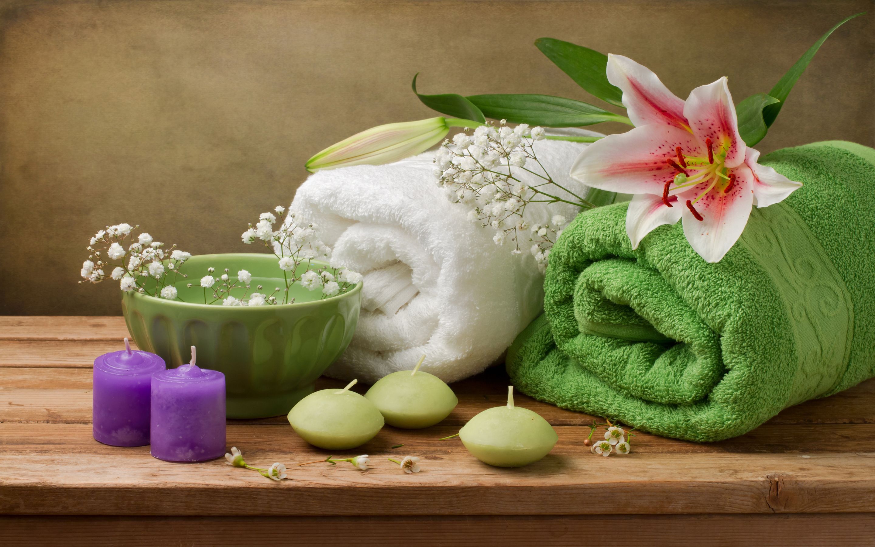 Una superficie de madera con una toalla verde, una toalla blanca, un cuenco verde con flores blancas y dos velas moradas.