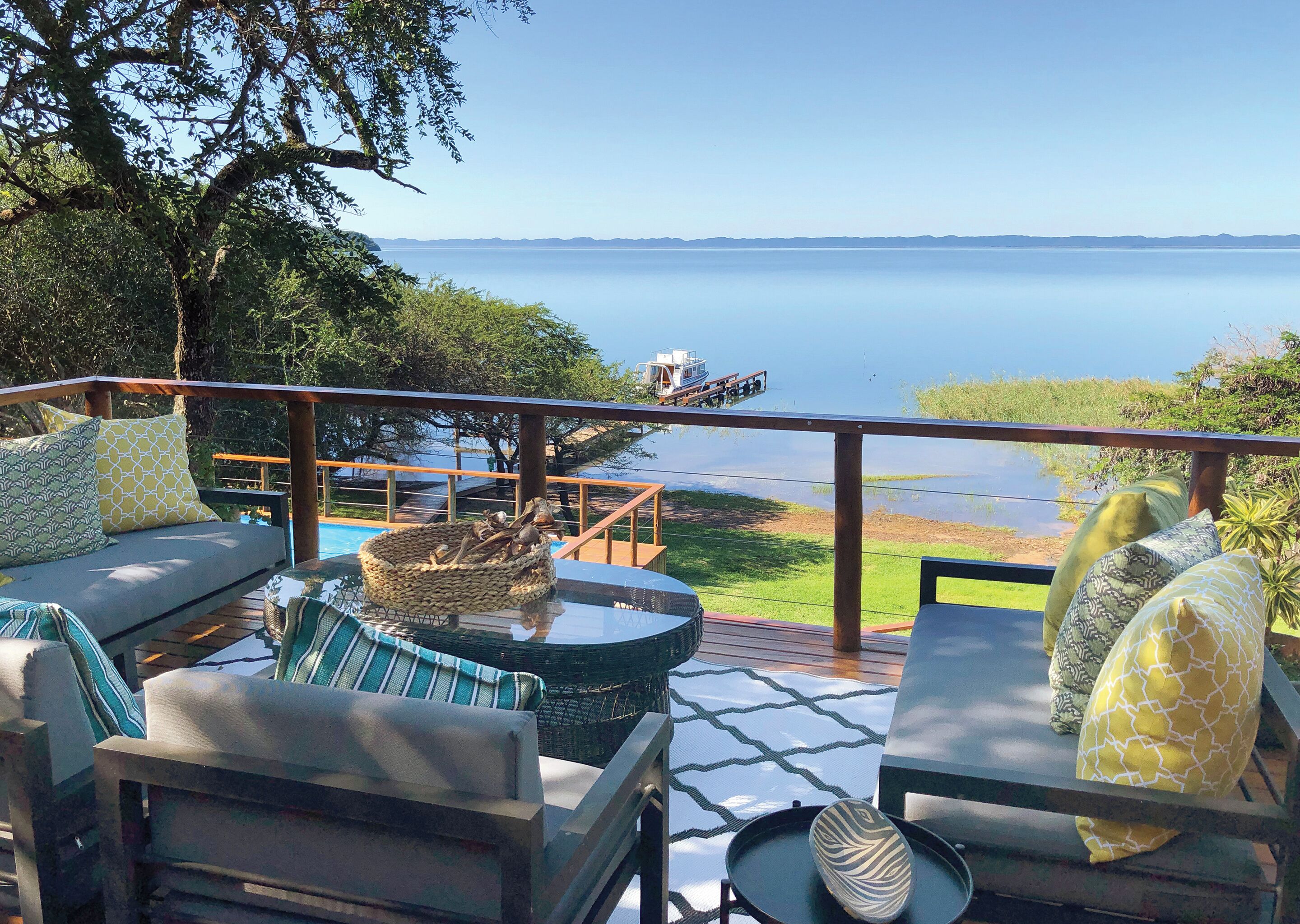 A deck with outdoor seating, a view of a calm lake, and a boat in the distance.