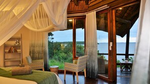 A bedroom with a large window, a bed, a chair, and a small table with a view of the outdoors. - Malkoha Lake St. Lucia (KwaNibela)