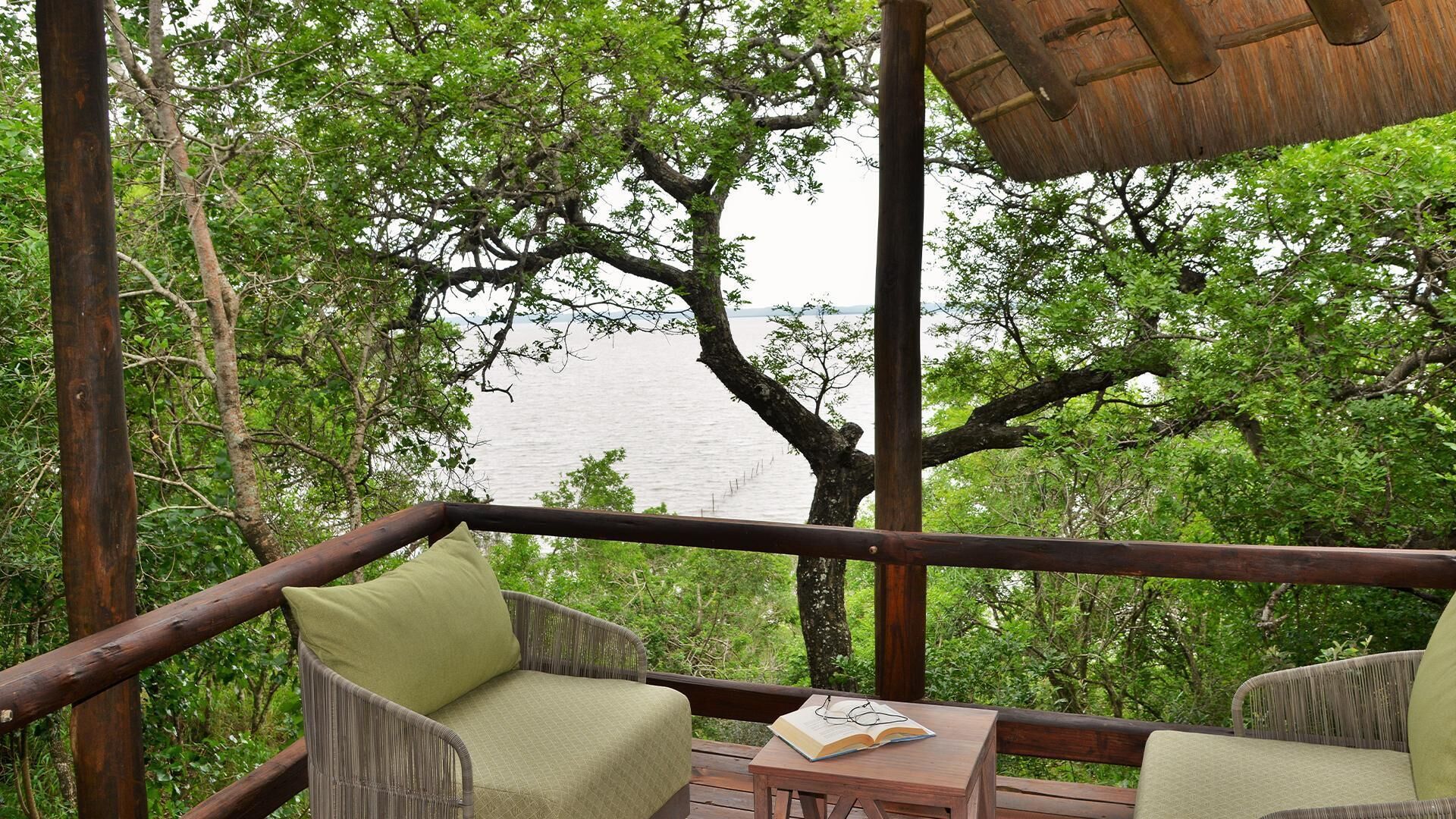 Une terrasse en bois, agrémentée de chaises et d’une table en osier, qui offre une vue sur une étendue d’eau et une végétation luxuriante.