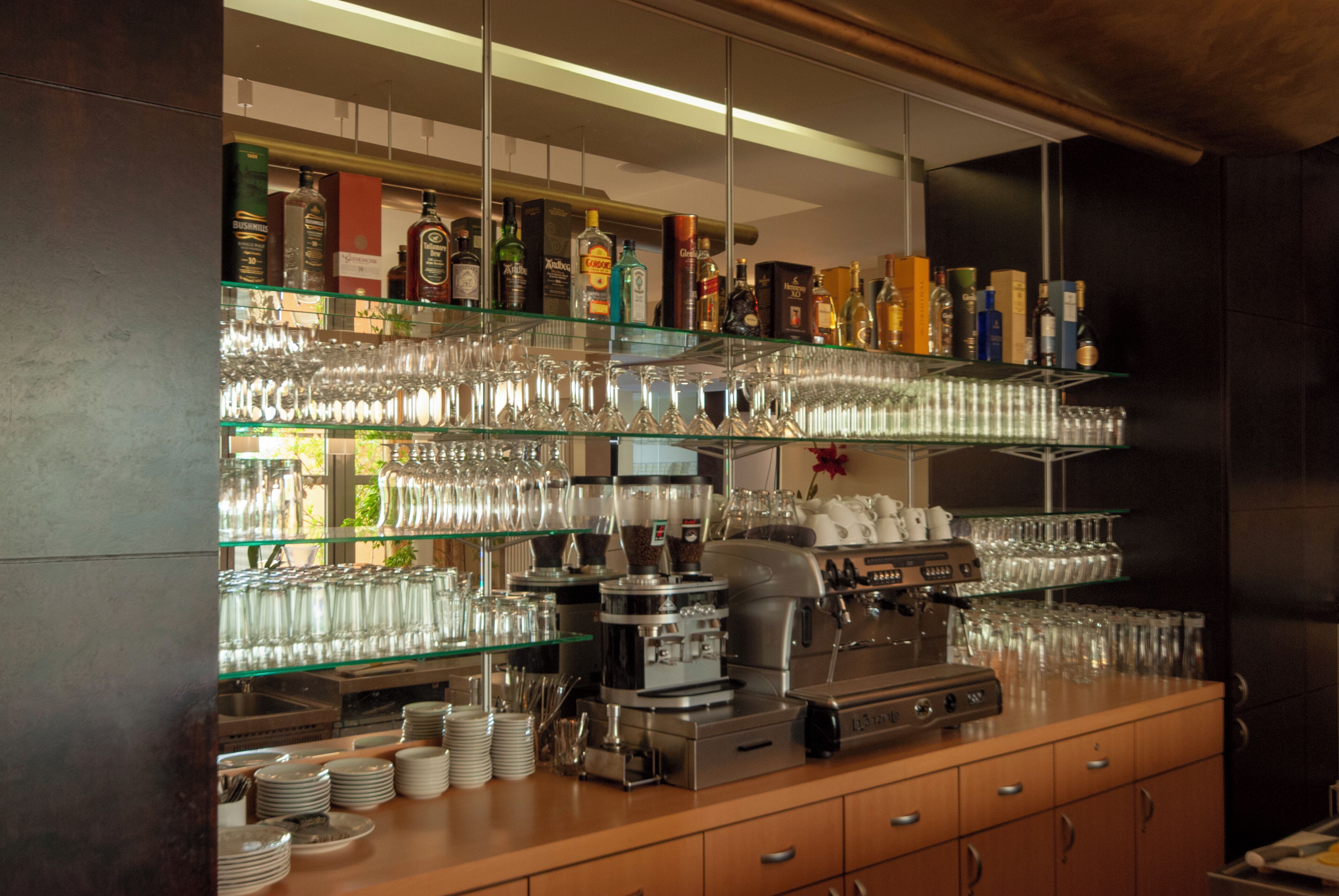 A bar area with shelves of glassware and bottles, a coffee machine, and stacked plates.