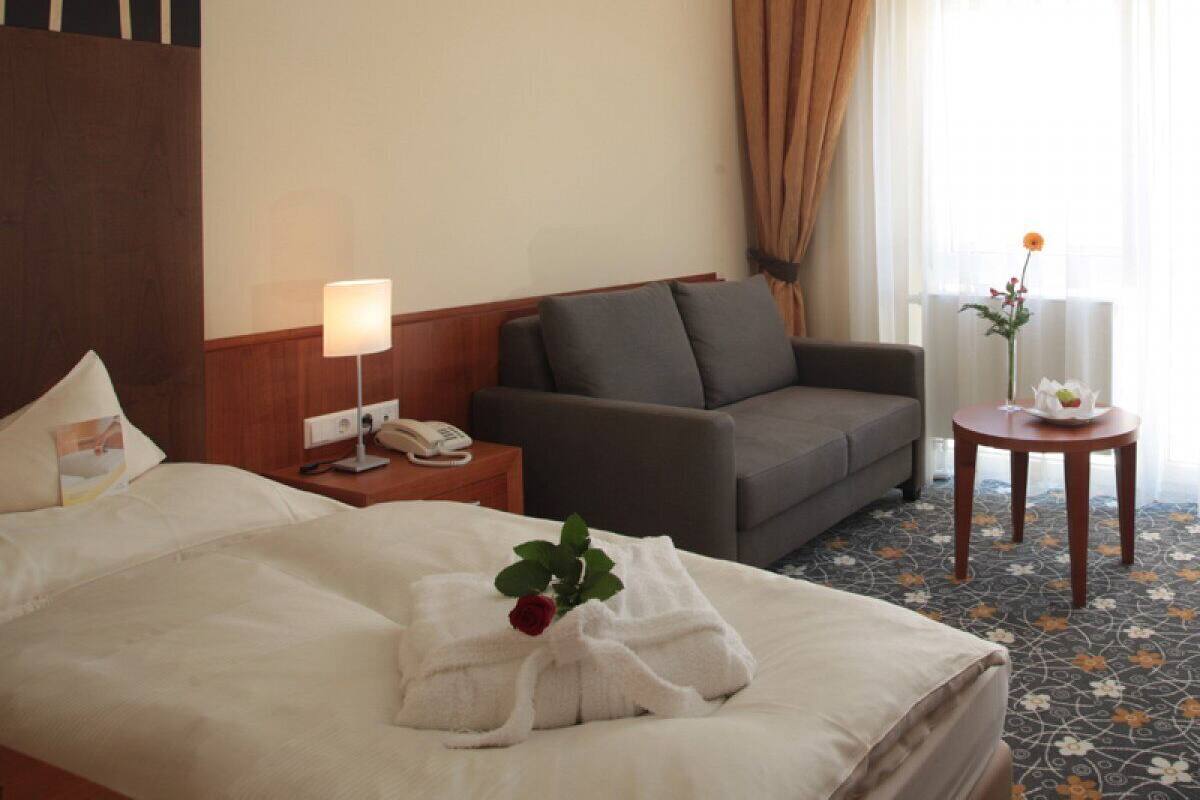 Habitación de hotel con cama, sofá, mesita de noche con lámpara, una pequeña mesa con un florero y una ventana con cortinas.