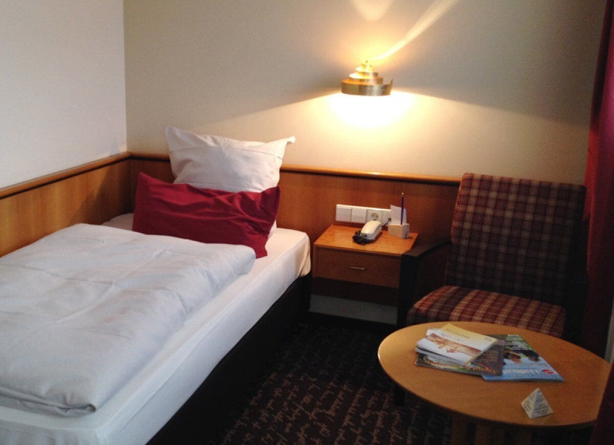 A hotel room with a bed, a nightstand with a lamp, a chair, and a small table with magazines.