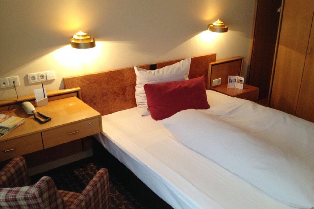 Habitación de hotel con una cama, dos mesitas de noche, un escritorio con teléfono y control remoto, y una silla.