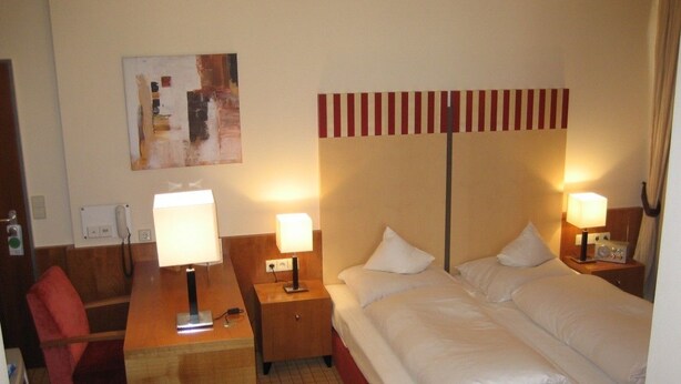 Habitación de hotel con cama, dos lámparas de noche, un escritorio con lámpara, una silla y un cuadro en la pared.