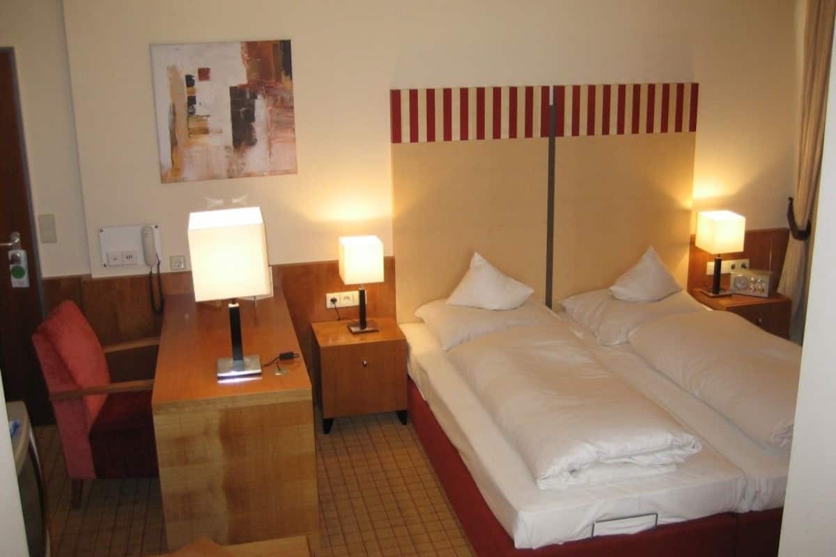 Habitación de hotel con cama, dos lámparas de noche, un escritorio con lámpara, una silla y un cuadro en la pared.