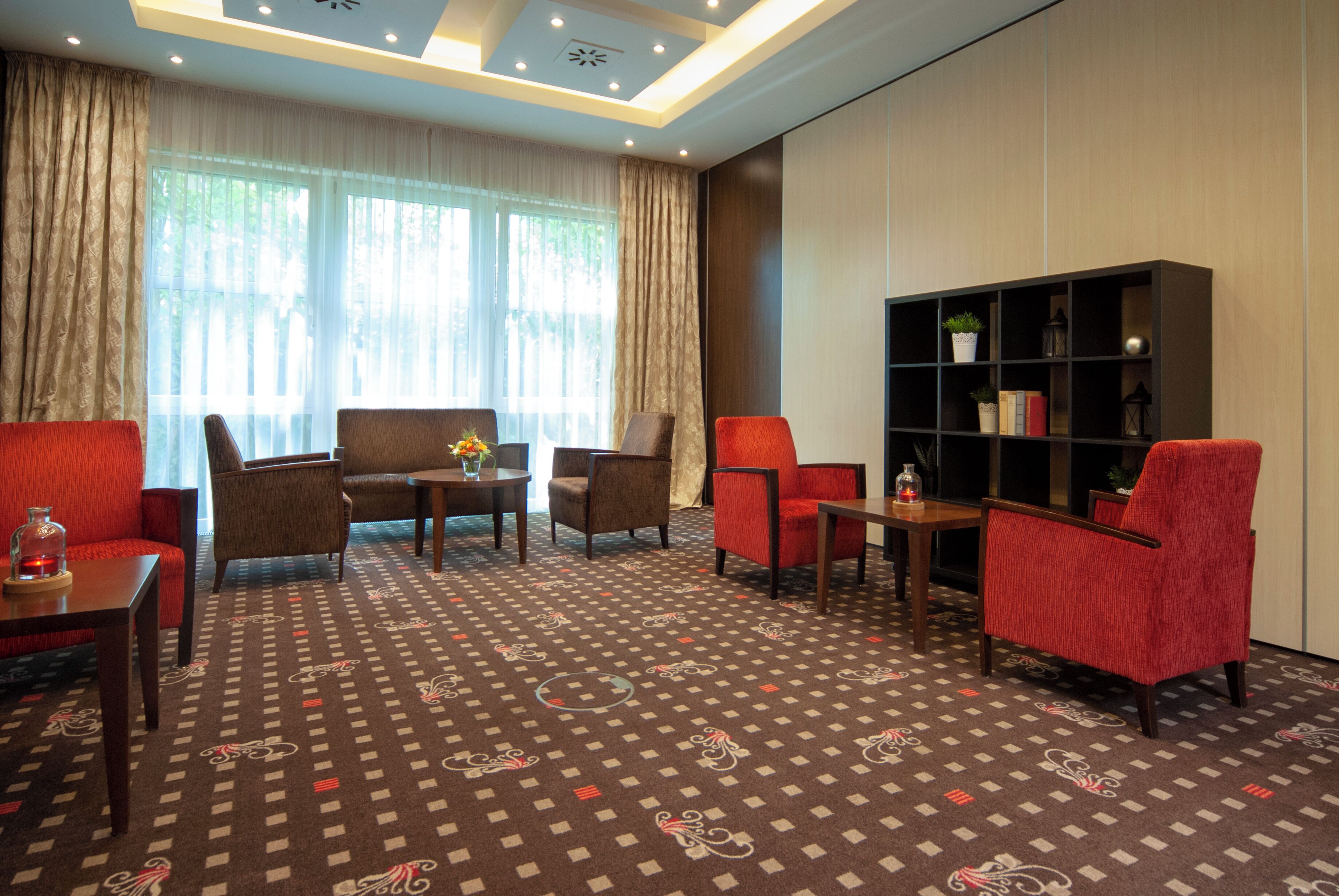A hotel room with a patterned carpet, a bookshelf, and a seating area with red and brown chairs.