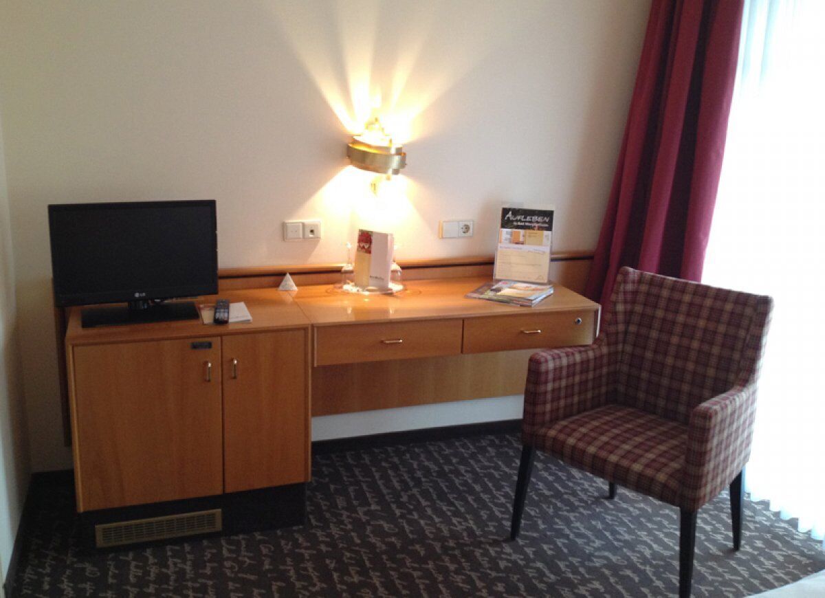 A hotel room with a wooden desk, a television, a lamp, a chair, and a carpeted floor.