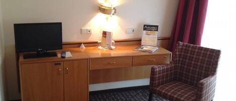 A hotel room with a wooden desk, a television, a lamp, a chair, and a carpeted floor.