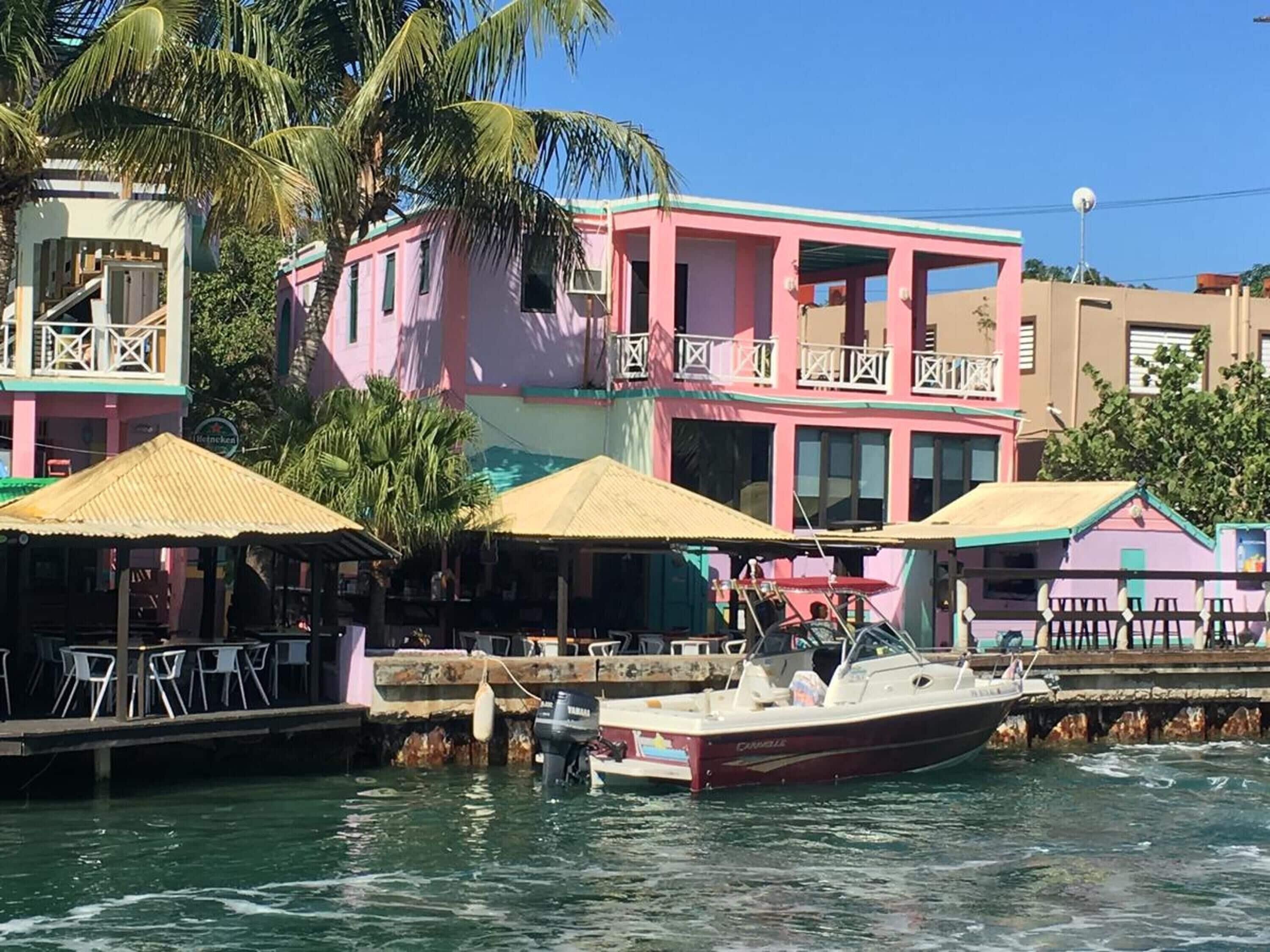 A waterfront building with a pink facade, a dock with a boat, and outdoor seating with umbrellas.