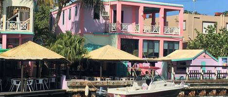 A waterfront building with a pink facade, a dock with a boat, and outdoor seating with umbrellas.