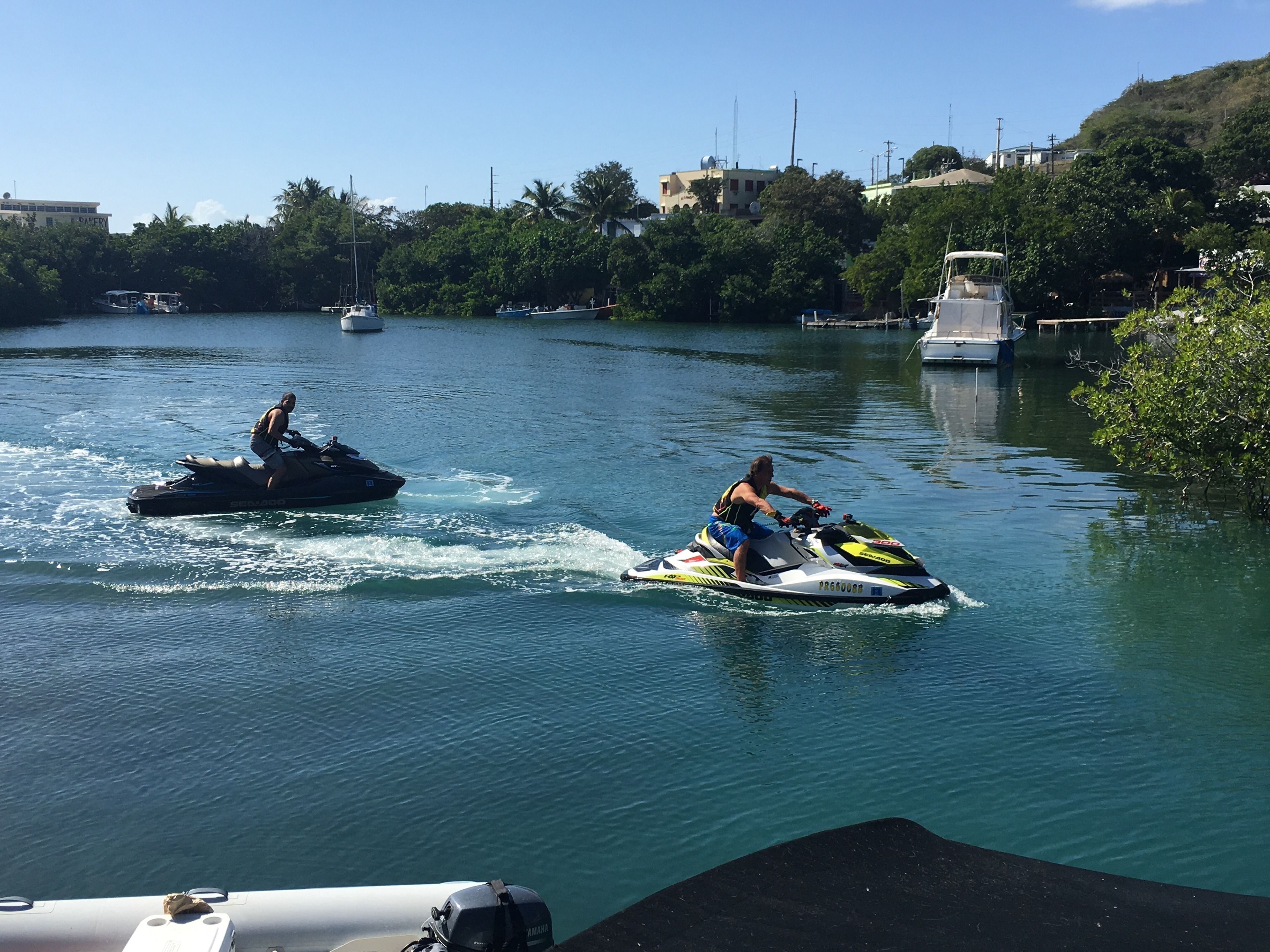 Two jet skis on a calm river with boats and greenery in the background.