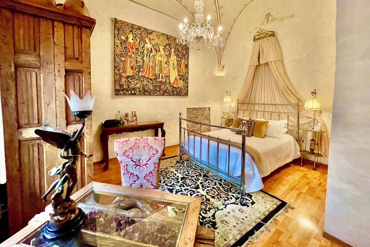 A bedroom with a canopy bed, a chandelier, a large floral painting, and a wooden cabinet.