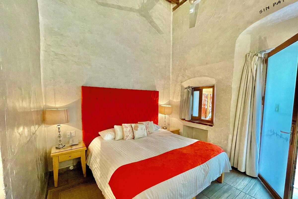 A bedroom with a red headboard, a bed with white bedding, a nightstand with a lamp, and a window with curtains.