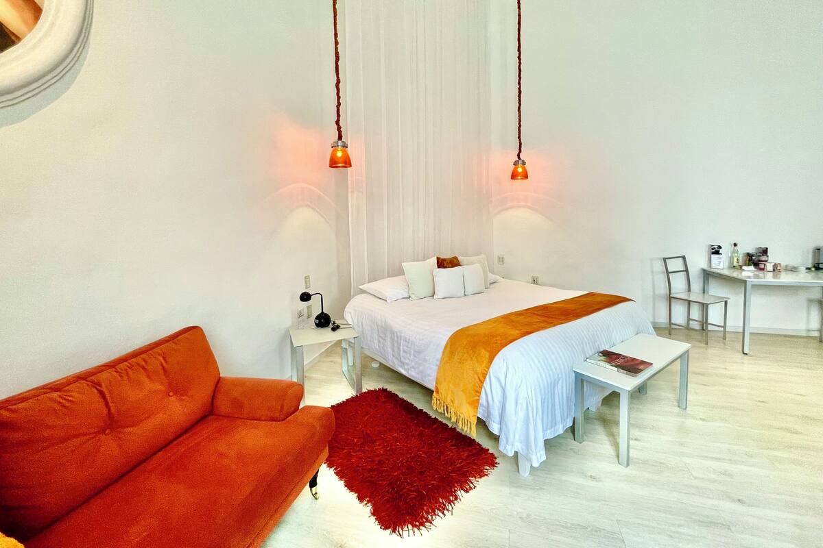 A modern hotel room with a bed, a small desk, a chair, and an orange sofa.