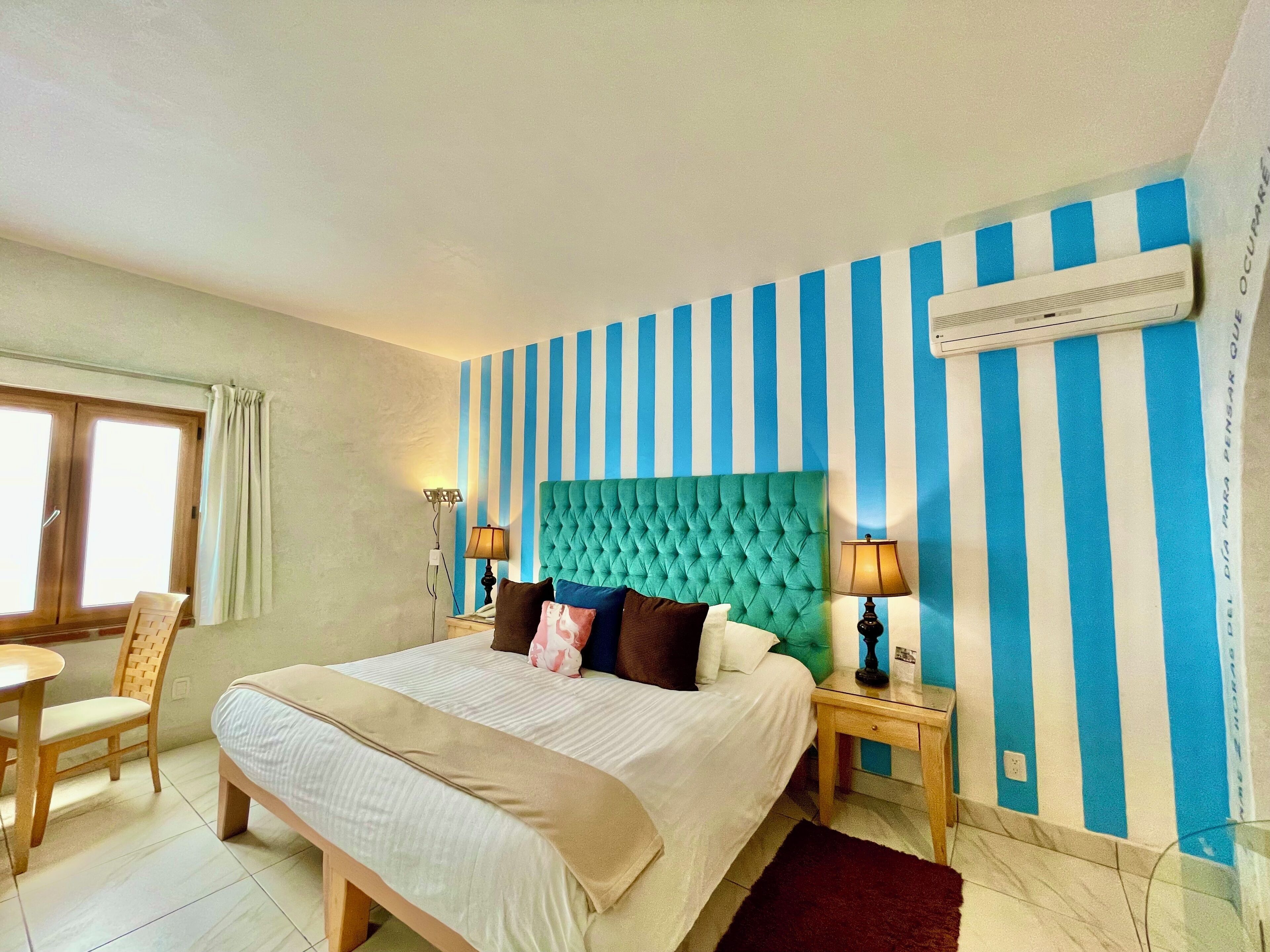 Habitación de hotel con una cama grande, dos mesitas de noche, un ventanal con cortinas y una pared a rayas.