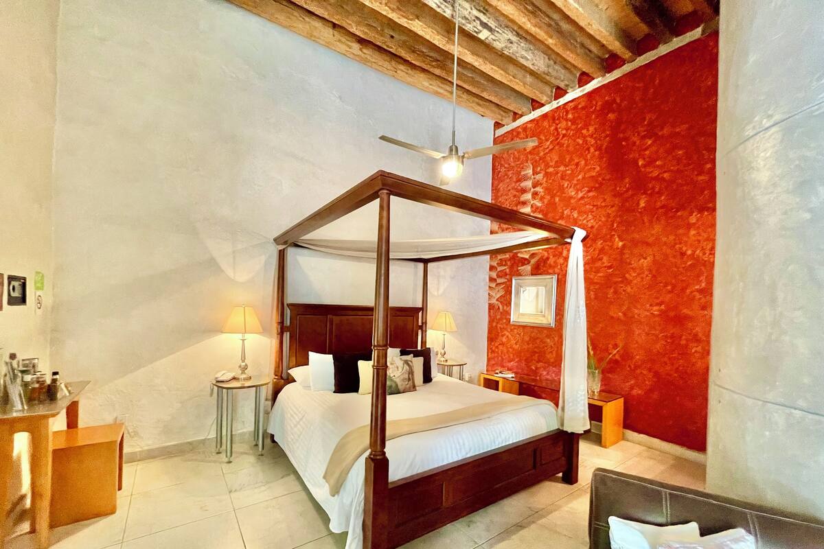 A four-poster bed with a wooden frame and white bedding, a ceiling fan, and a red accent wall.