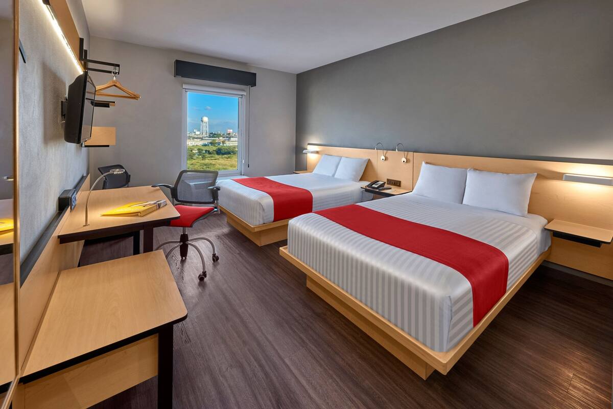 A hotel room with two beds, a desk, a chair, a TV mounted on the wall, and a window with a city view.
