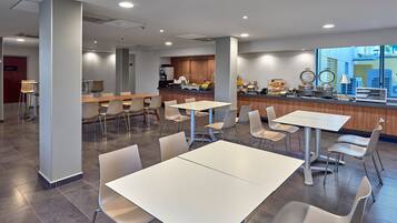 A modern dining area with white tables and chairs, a kitchen counter with food items, and a view of the outside.