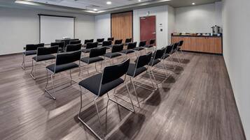 Ein Konferenzraum mit Reihen schwarzer Stühle, einer Projektionsleinwand, einer Holztür und einer Küchenzeile.