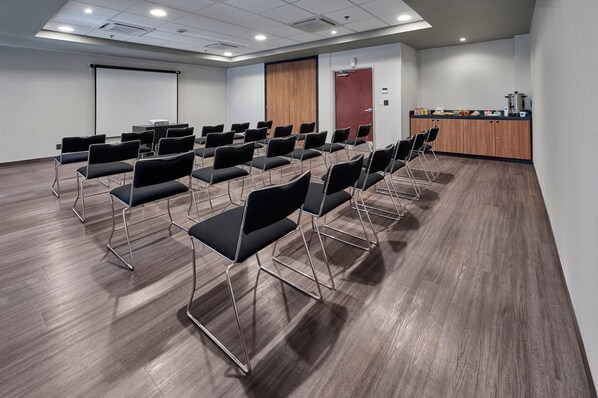 A conference room with rows of black chairs, a projector screen, a wooden door, and a kitchenette area.