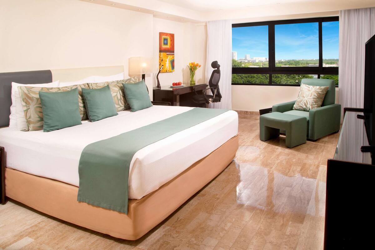 Habitación de hotel con una cama grande, un escritorio, una silla y vistas a la ciudad a través de amplios ventanales.