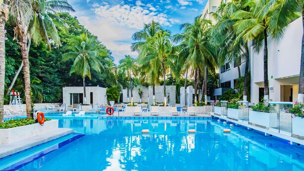 A swimming pool with lounge chairs and palm trees, surrounded by a white building with multiple floors. - Smart Cancun The Urban Oasis (Cancun)