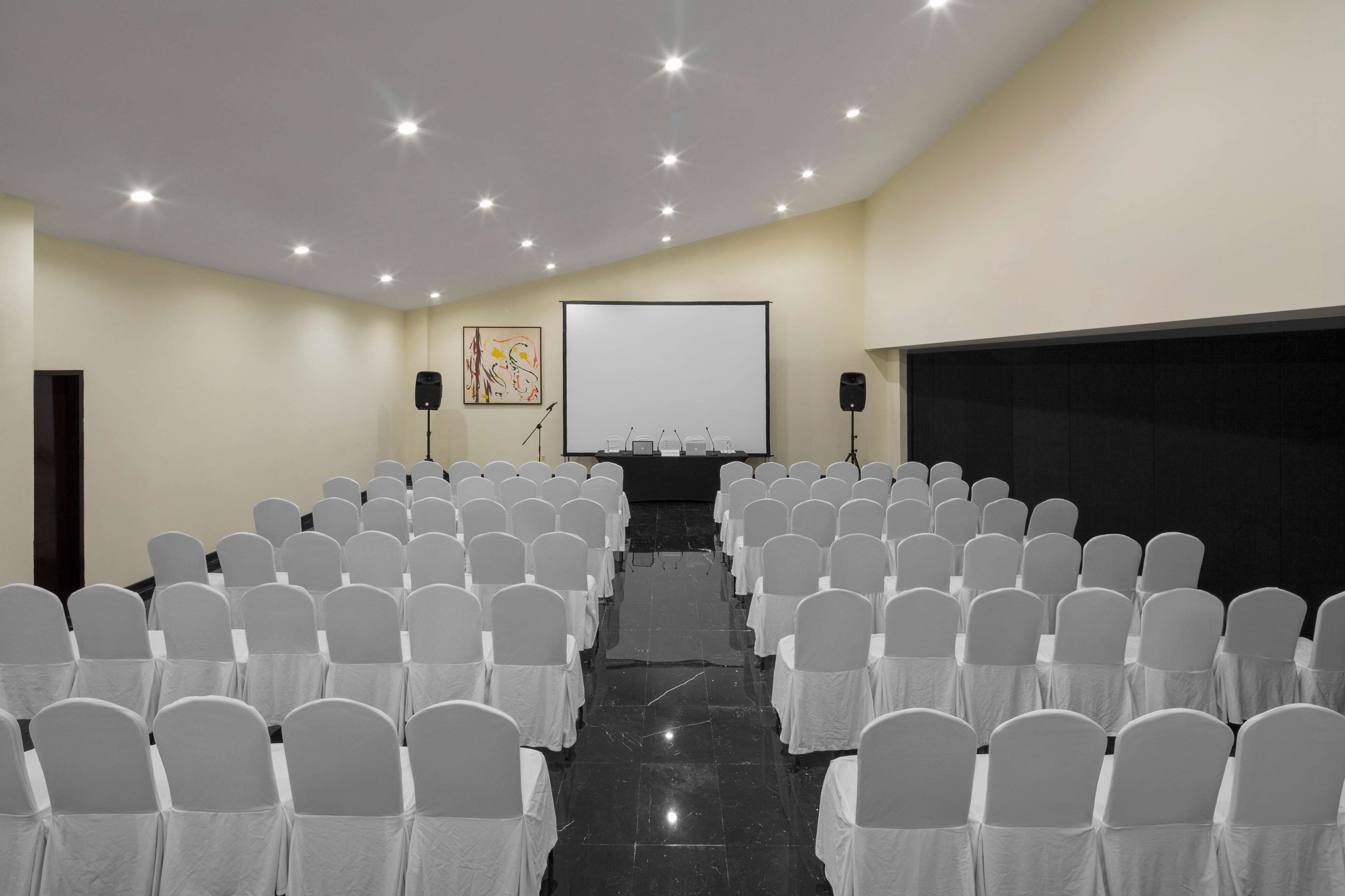 A conference room with rows of white chairs, a projector screen, and a speaker on stands.