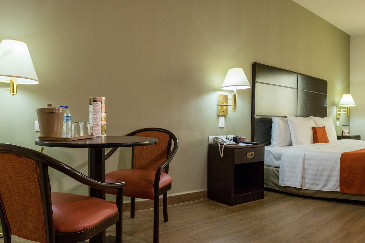 A hotel room with a bed, a desk with chairs, a bedside table with a lamp, a window, and an air conditioning unit.