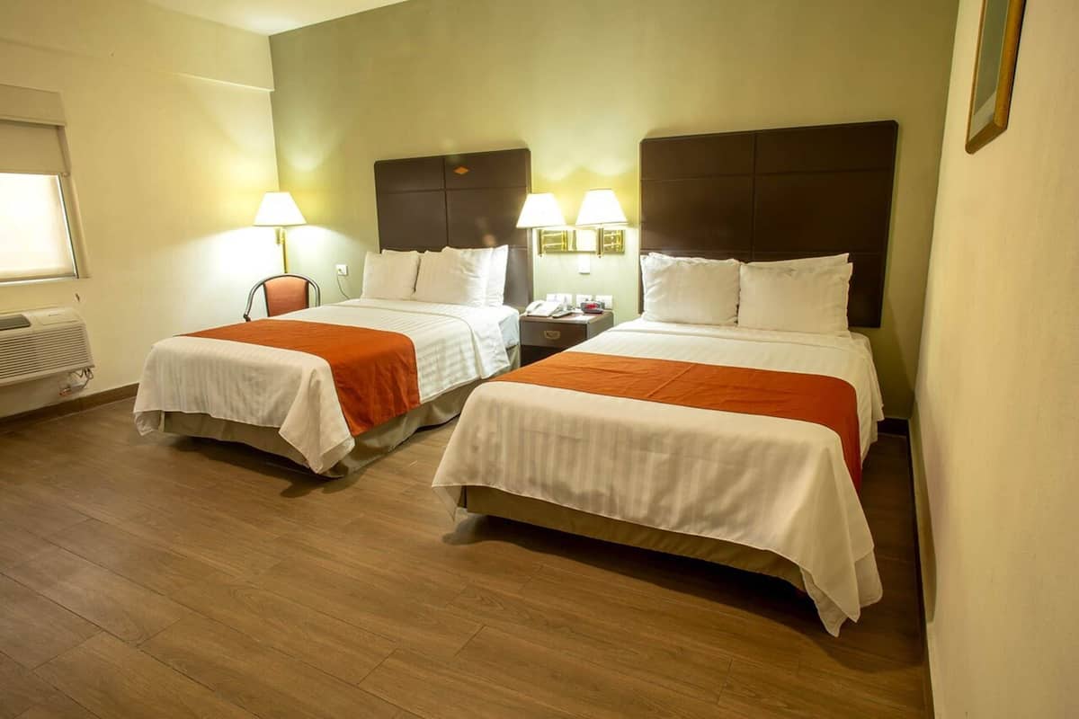 A hotel room with two beds, each with a white bedspread and orange accents, a nightstand with a lamp, and a window with blinds.