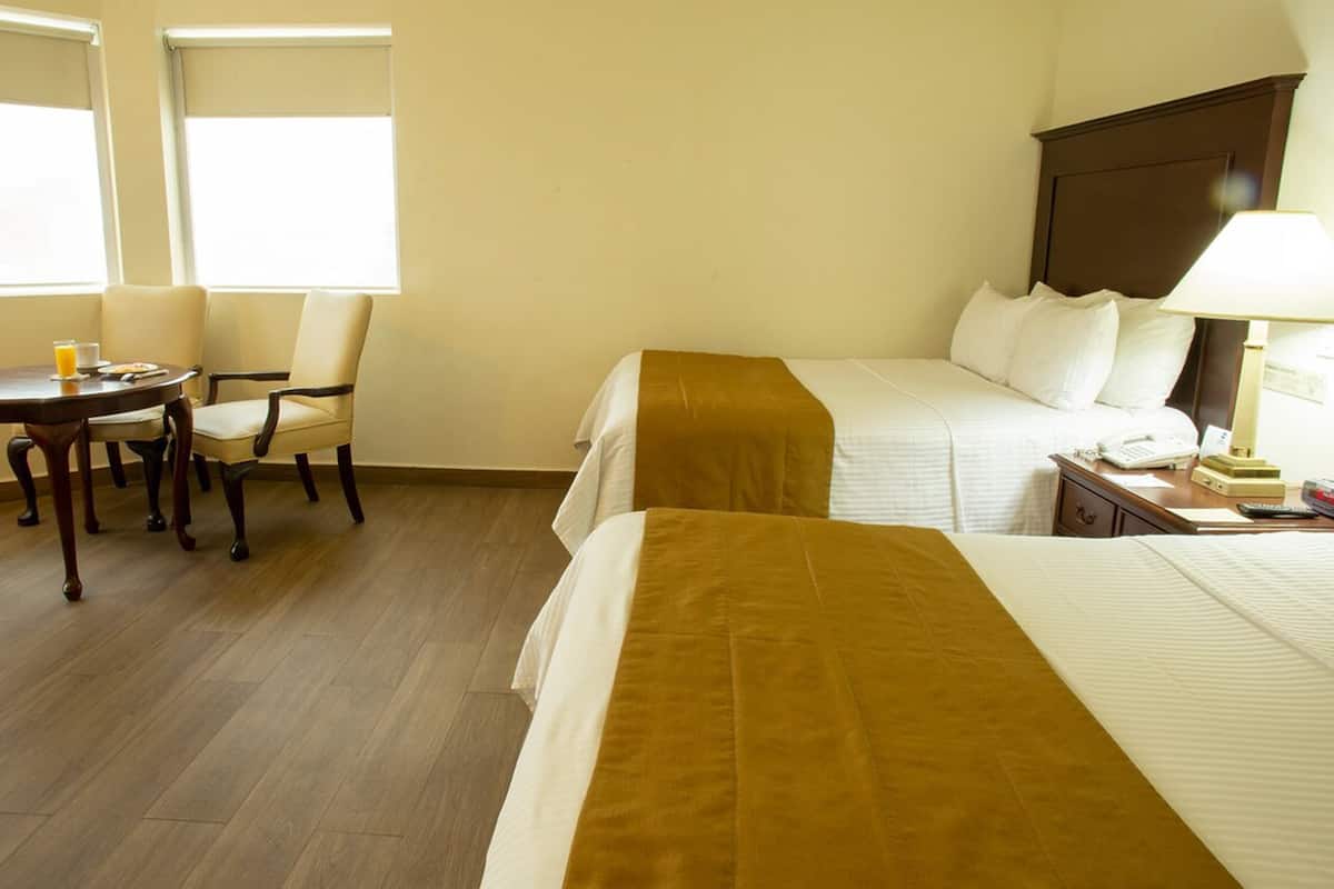 A hotel room with two beds, a small table with a chair, a bedside table with a lamp, and a window with blinds.