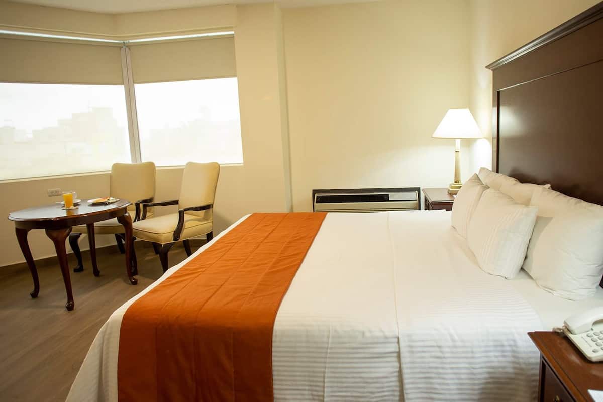 A hotel room with a large bed, a desk with a lamp, two chairs, and a window with blinds.