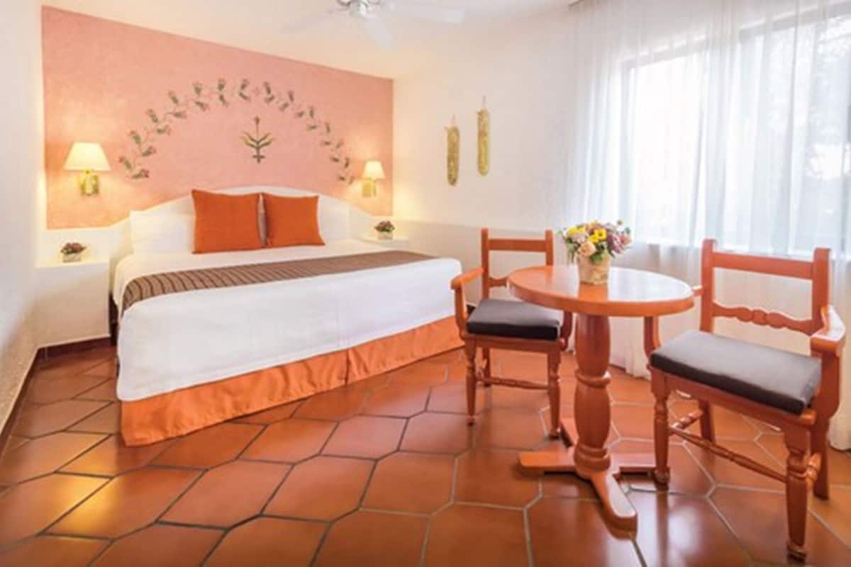 A hotel room with a bed, two chairs, a small table, a vase with flowers, and a wall with a floral design.