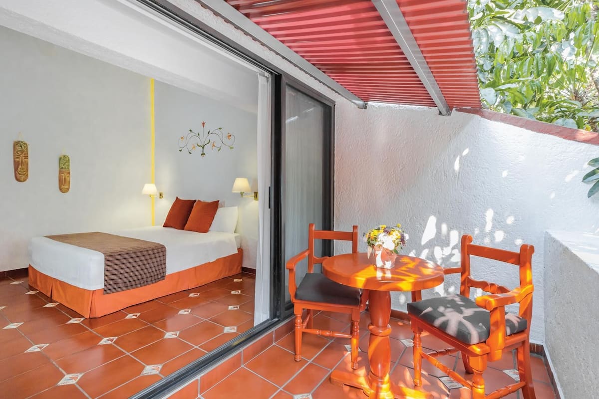 A room with a bed, a table with chairs, a small balcony with a red awning, and a window with a view of greenery.