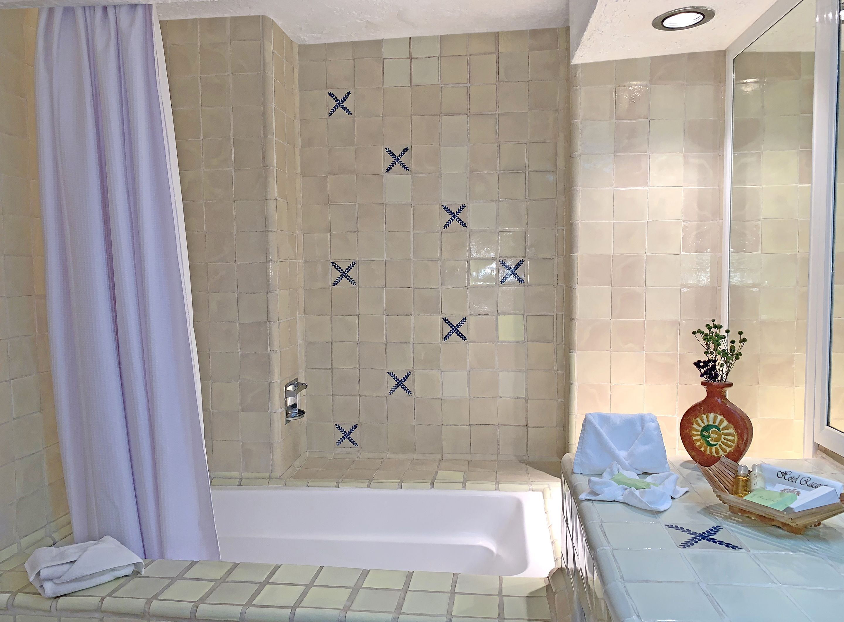 Uma casa de banho com banheira, zona de duche com azulejos decorativos, toucador com artigos de higiene e um vaso com flores.