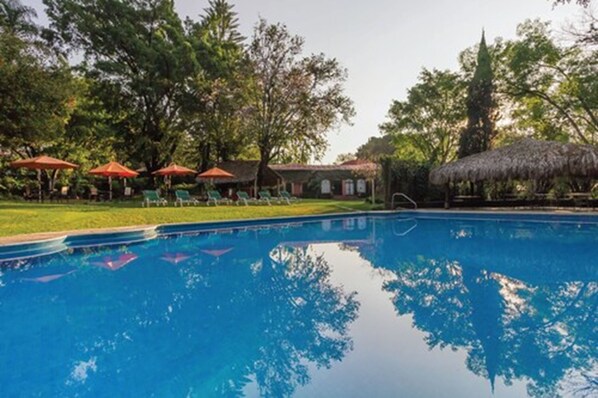Una piscina con sillas de descanso y sombrillas, rodeada de árboles y una estructura con techo de paja.