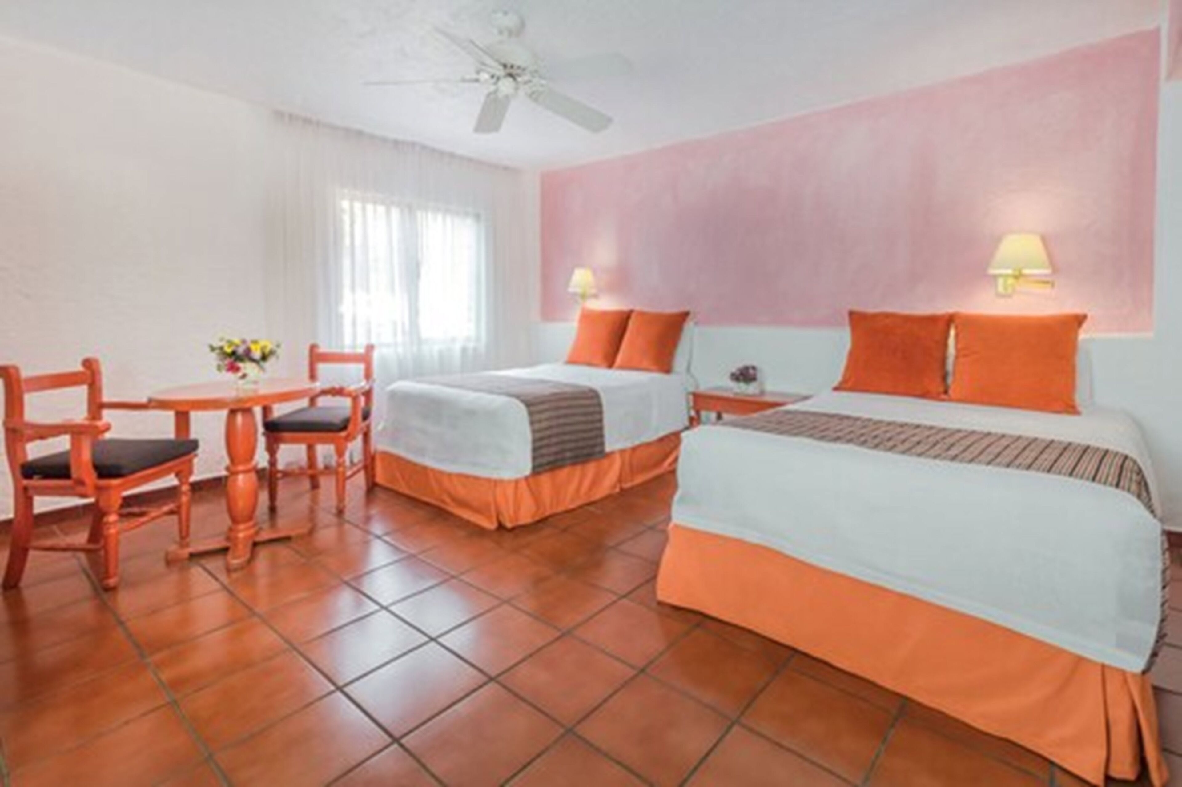 A hotel room with two beds, a dining table with chairs, and a ceiling fan.