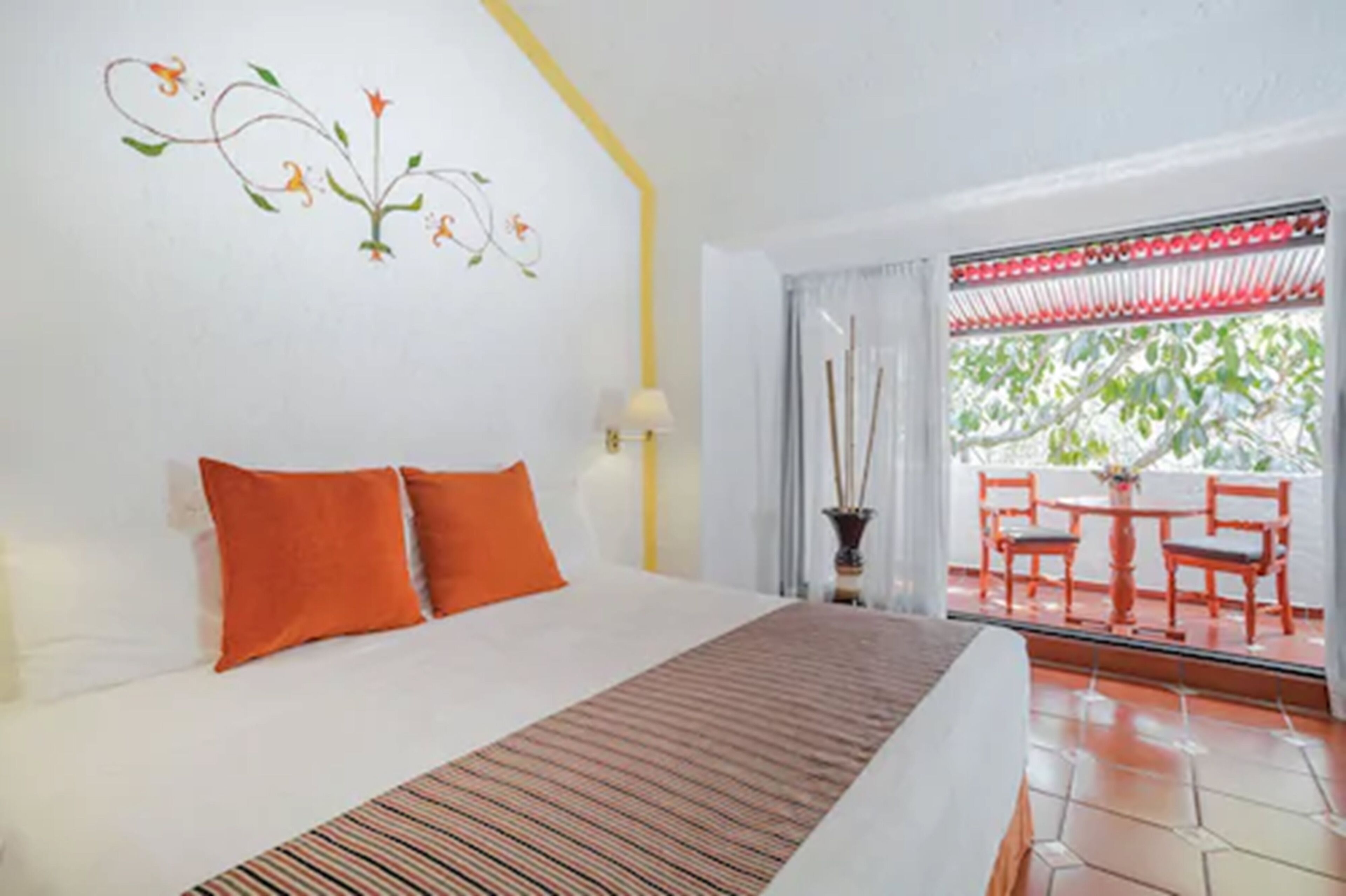 A hotel room with a bed, orange pillows, a striped area rug, a wall decoration, a window with a view of the outdoors, and a table with chairs outside.