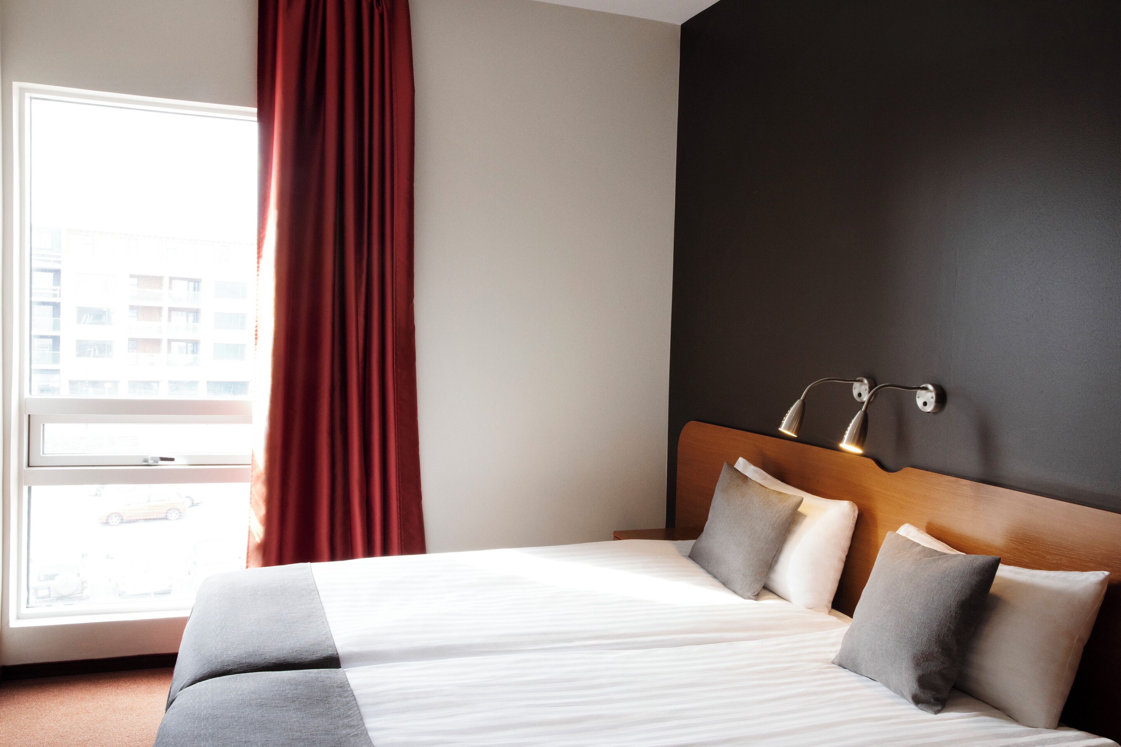 Une chambre d’hôtel avec un grand lit, une fenêtre donnant sur des immeubles et un mur sombre agrémenté d’un luminaire.