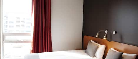 Une chambre d’hôtel avec un grand lit, une fenêtre donnant sur des immeubles et un mur sombre agrémenté d’un luminaire.