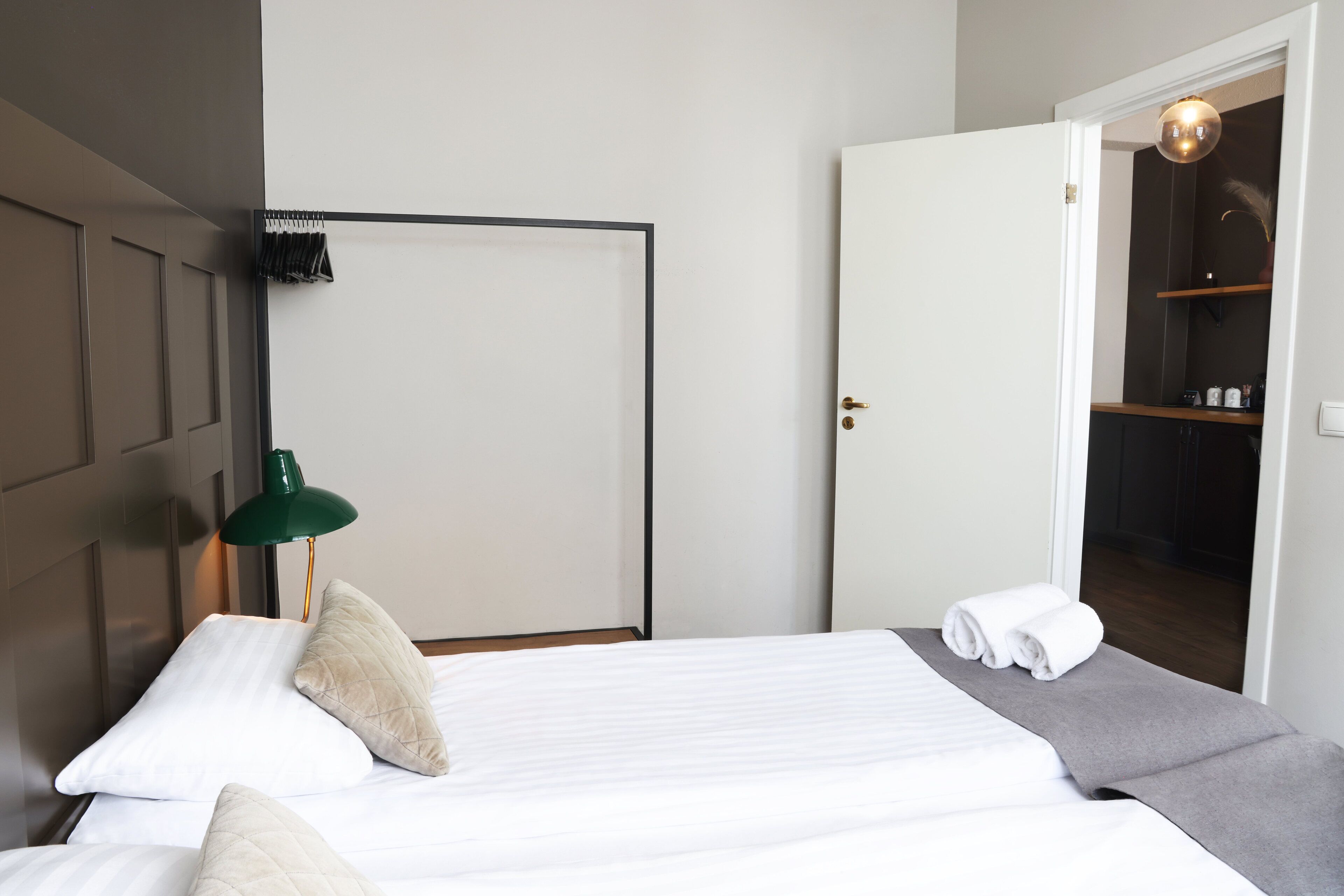 Ein ordentlich bezogenes Bett mit Kissen, eine grüne Schreibtischlampe und ein Kleiderschrank mit Spiegel.