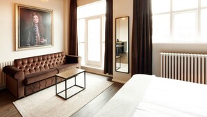 A modern hotel room with a large bed, a brown leather sofa, a small wooden table, a framed portrait on the wall, and a large window with curtains. - Fosshotel Barón (Reykjavik)