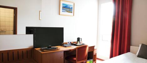 A hotel room with a bed, a desk with a computer, a chair, a window with red curtains, and a framed picture on the wall.