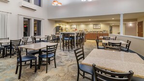 Une salle à manger d’hôtel avec plusieurs tables et chaises, une cuisine en arrière-plan et des luminaires au plafond.