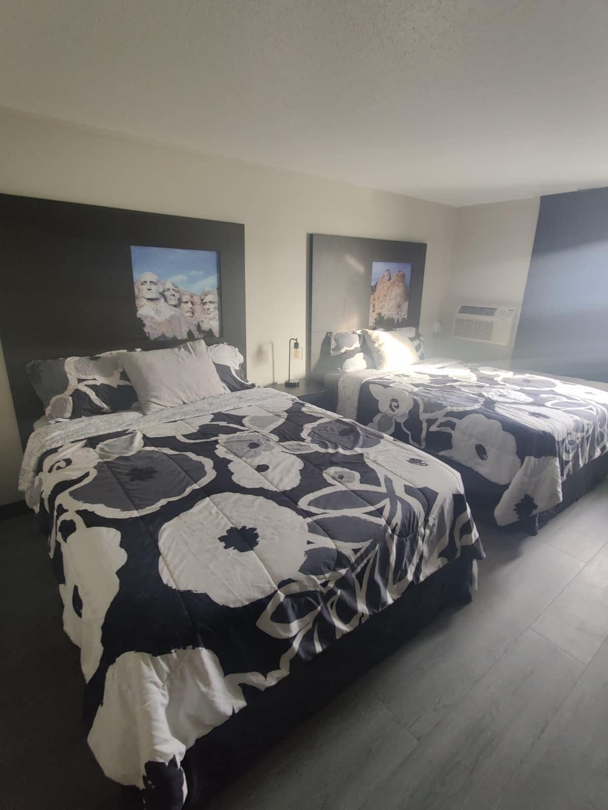 A hotel room with two beds, each with a floral-patterned bedspread, a nightstand, a lamp, and wall art depicting mountain landscapes.